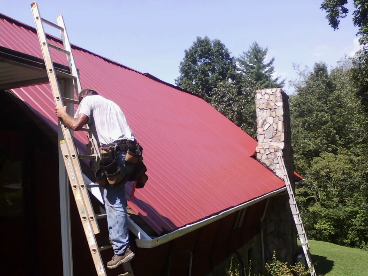Roofer on a ladder beside a red metal roof and stone chimney — Soffit & Fascia Repair by Roofing Brothers in Two Dot